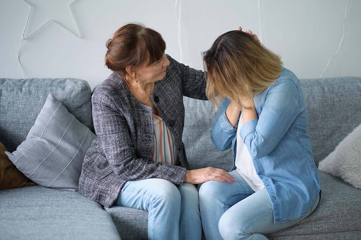 5 Signs of Alcoholism in a Loved One Woman on couch consoling friend about signs of alcoholism in a loved one