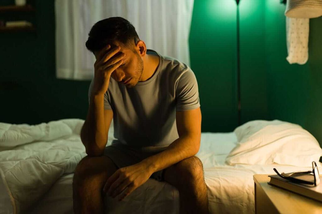 Person sitting at the edge of the bed late at night with a headache while suffering from symptoms of alcohol poisoning